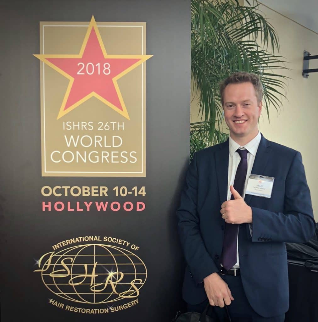 A smiling man in a suit and tie giving a thumbs up next to a promotional banner for the "2018 ISHRS 26th World Congress on Hair Replacement" in Hollywood.