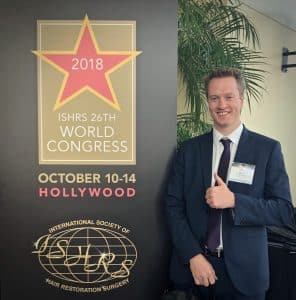 A smiling man in a suit and tie giving a thumbs up next to a promotional banner for the "2018 ISHRS 26th World Congress on Hair Replacement" in Hollywood.