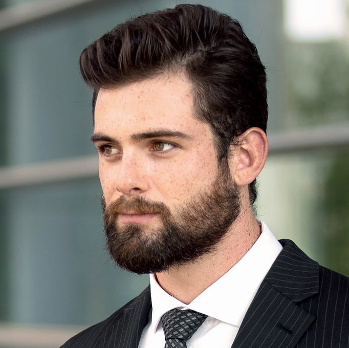 A portrait of a man with hair regrowth, displaying a full beard and thick, neatly groomed hair. He's wearing a pinstripe suit and a dark tie, looking to the side with
