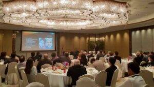 A conference room with attendees seated at round tables, listening to a speaker presenting near a large screen displaying images and text about hair implant surgery, under elegant chandelier lighting.