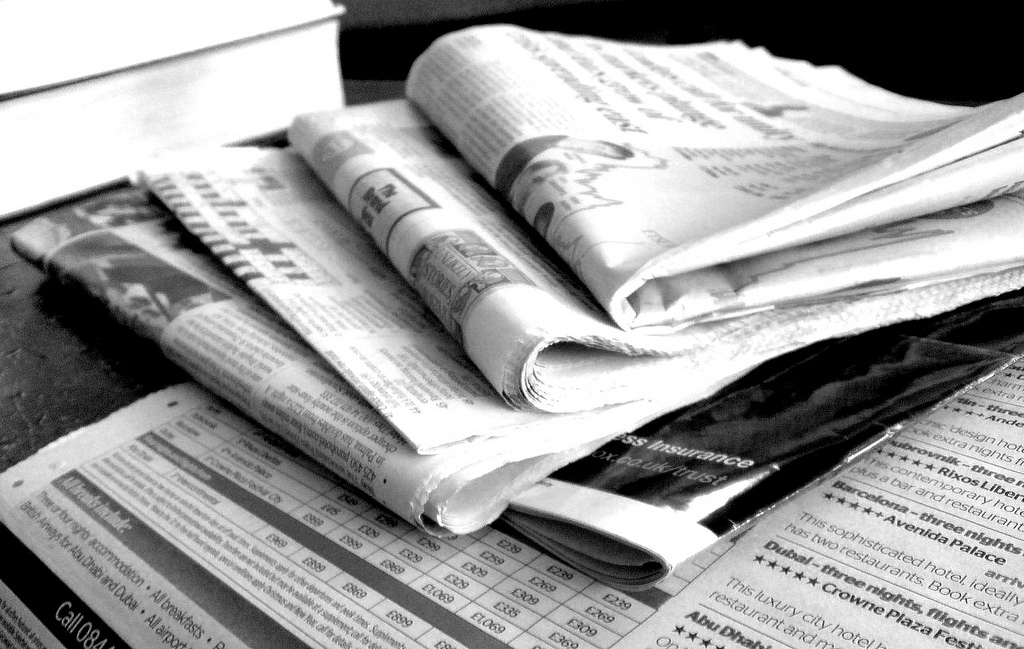 A black and white image of a pile of folded and rolled-up newspapers, with visible text and images on the pages, including an article about hair regrowth.