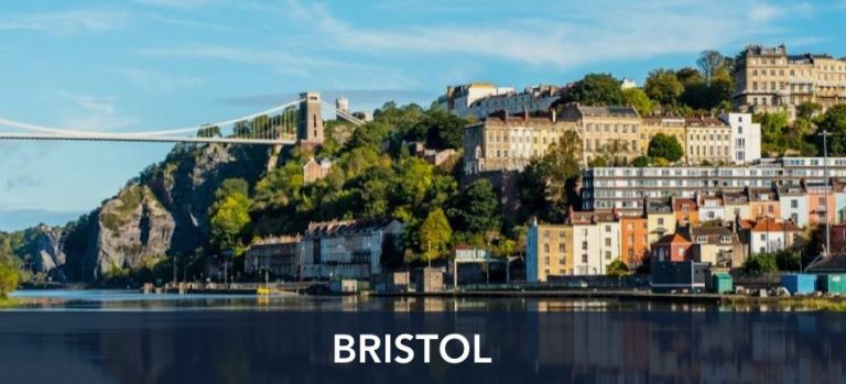 A panoramic view of Bristol featuring colorful houses on the hillside, green vegetation, and the Clifton Suspension Bridge, with a calm river in the foreground.