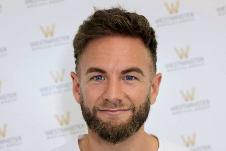 A man with a beard and short, styled hair implant smiles gently at the camera against a backdrop with a repeating logo pattern. He wears a white shirt, and the lighting is bright and even.