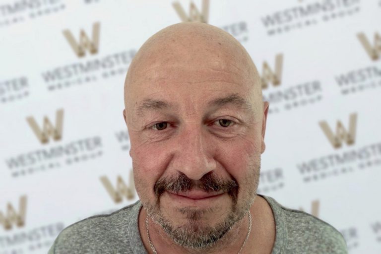 Close-up of a smiling man with male pattern baldness and a goatee, wearing a gray shirt. He stands in front of a backdrop marked with repeated "Westminster" logos.