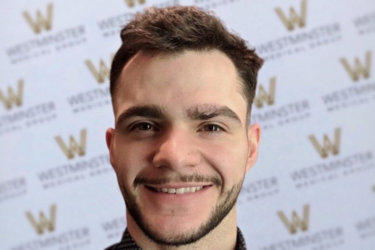 A young man smiling, with short dark hair potentially benefiting from hair regrowth treatment and a beard, standing in front of a backdrop with the repeated "Westminster" logo. He is wearing a check