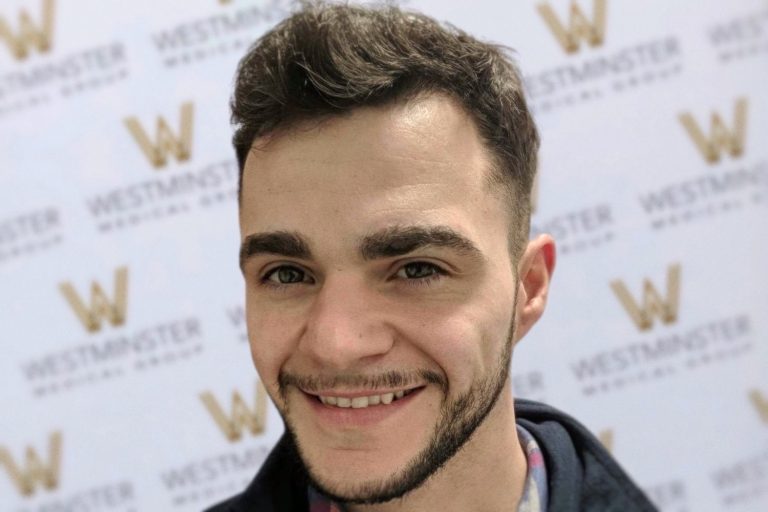 A young man with signs of hair regrowth and a light beard smiling in front of a backdrop with "Westminster" logos repeated in a lattice pattern.