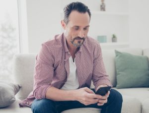 A man with a beard, wearing a red checkered shirt, sitting on a white couch and using a smartphone in a well-lit living room with green cushions, has undergone hair regrowth treatment.