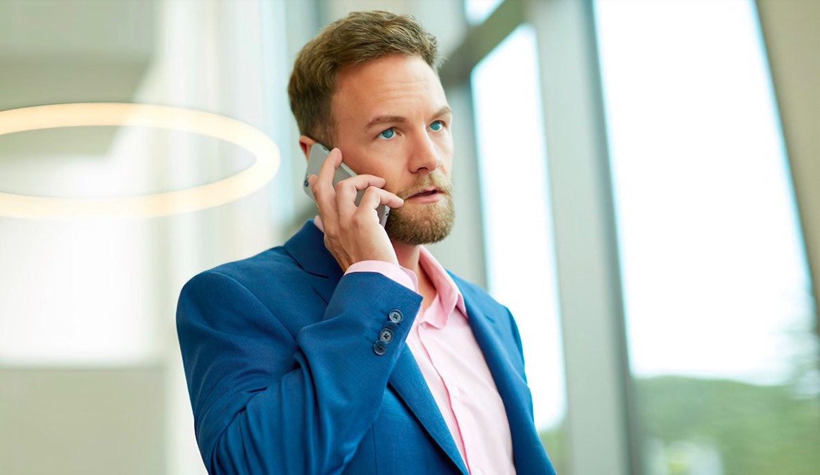 A professional man in a blue suit and pink shirt speaking on a mobile phone, standing in a brightly lit modern office with large windows and a circular light fixture overhead, discussing female hair loss treatments.