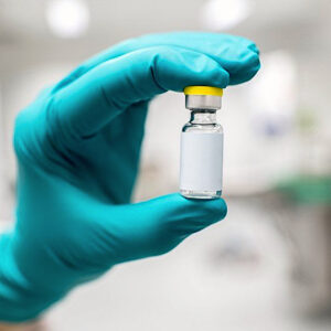 A hand wearing a teal glove holds a small vaccine vial in a laboratory setting, indicating a focus on medical or scientific research related to female hair loss.