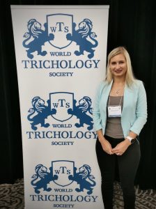 A woman with blond hair, wearing a teal blazer and black pants, stands smiling in front of a banner for the world trichology society addressing female hair loss, which features blue and white colors