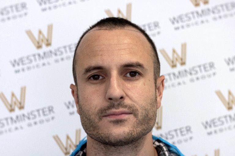 A man with a stubble beard and signs of male pattern baldness stands in front of a backdrop with the Westminster Woods logo, looking directly at the camera.