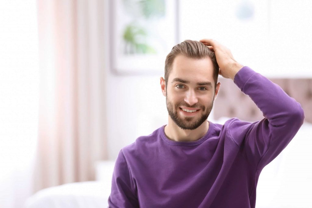 A young man with a beard and a smile, wearing a purple sweater, sits in a brightly lit room with white walls and small framed artworks on the wall. He is casually touching his hair replacement with