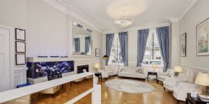 Elegant living room with high ceilings, white walls, a large aquarium, classic fireplace, and plush white sofas. Large windows draped with blue curtains add a touch of color.