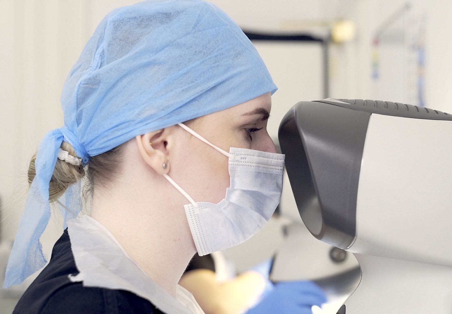 A female medical professional, wearing a surgical cap and mask, intently looks into a large diagnostic machine in a bright clinical setting, specializing in female hair loss.
