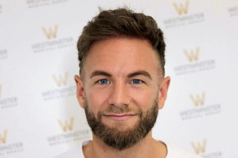 A man with a well-groomed beard and short hair implant, smiling at the camera. He stands in front of a white background with a subtle logo pattern.