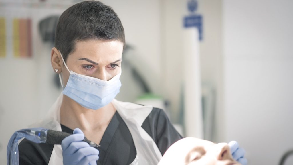 A focused female dentist wearing a surgical mask and protective gloves examines a patient's oral health using dental instruments in a well-lit clinic, specializing in treatments for conditions like male pattern baldness.