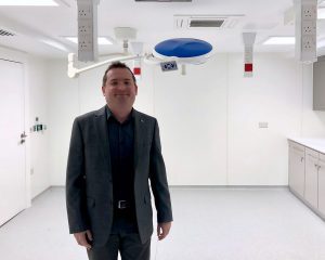 A man in a suit smiling in a modern medical or dental office with bright lighting and a large overhead surgical lamp. The room appears clean and well-equipped for hair transplant services.