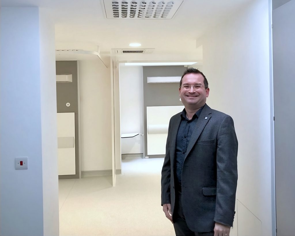 A smiling man with freshly restored hair in a dark blazer stands in a well-lit, modern office corridor with white walls and open doorways leading to brightly lit rooms.