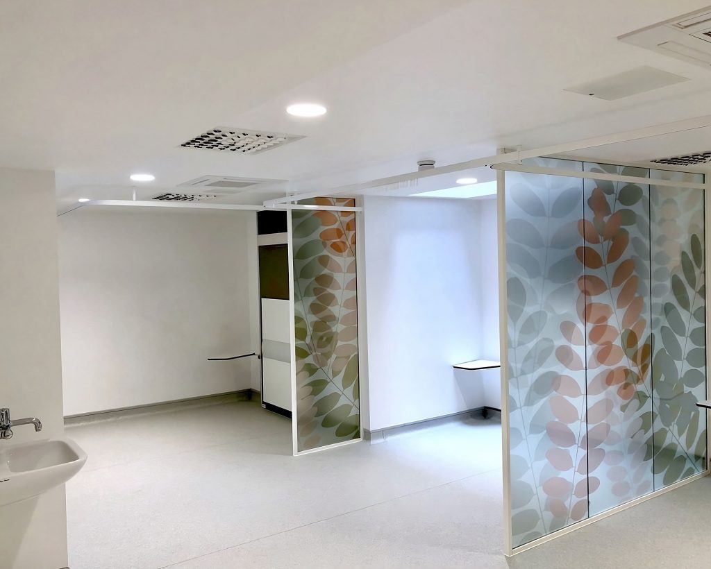 Modern office restroom interior with frosted glass doors featuring a leaf design, white walls, and a tiled floor. A sink is visible on the left side. This restroom also includes informational posters about male pattern
