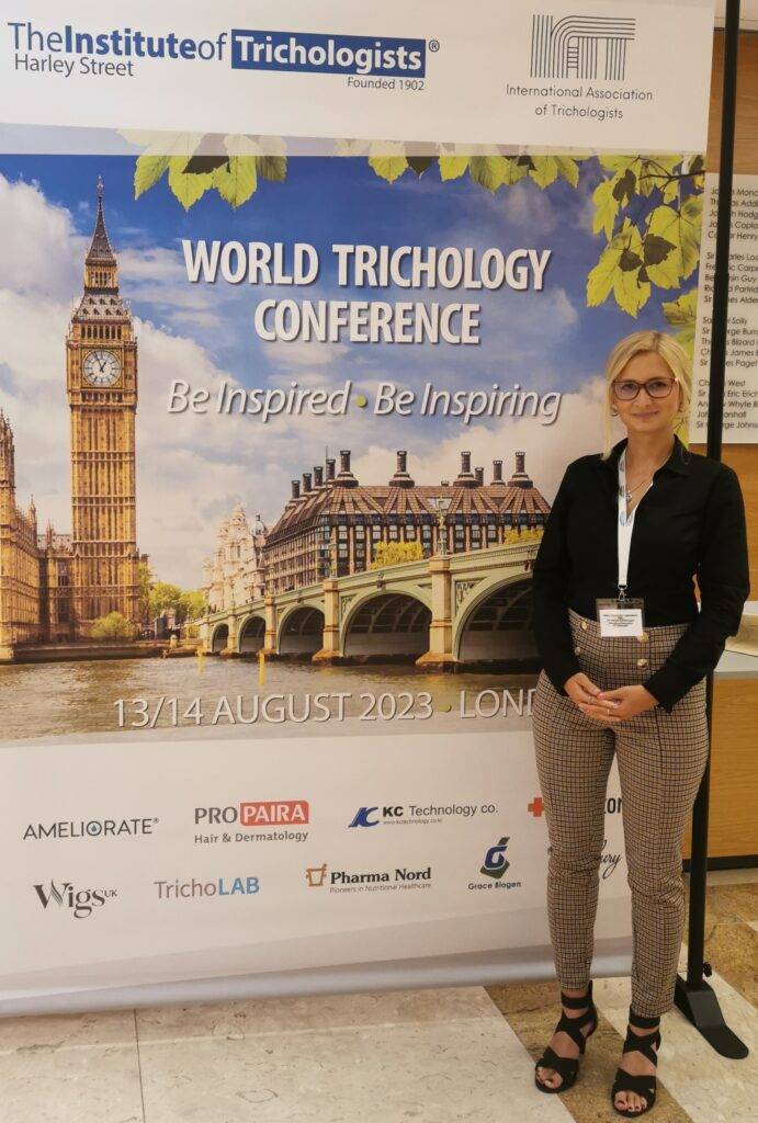 A woman posing in front of a banner for the "world trichology conference" held on August 13/14, 2023, in London. The banner features images of Big Ben and