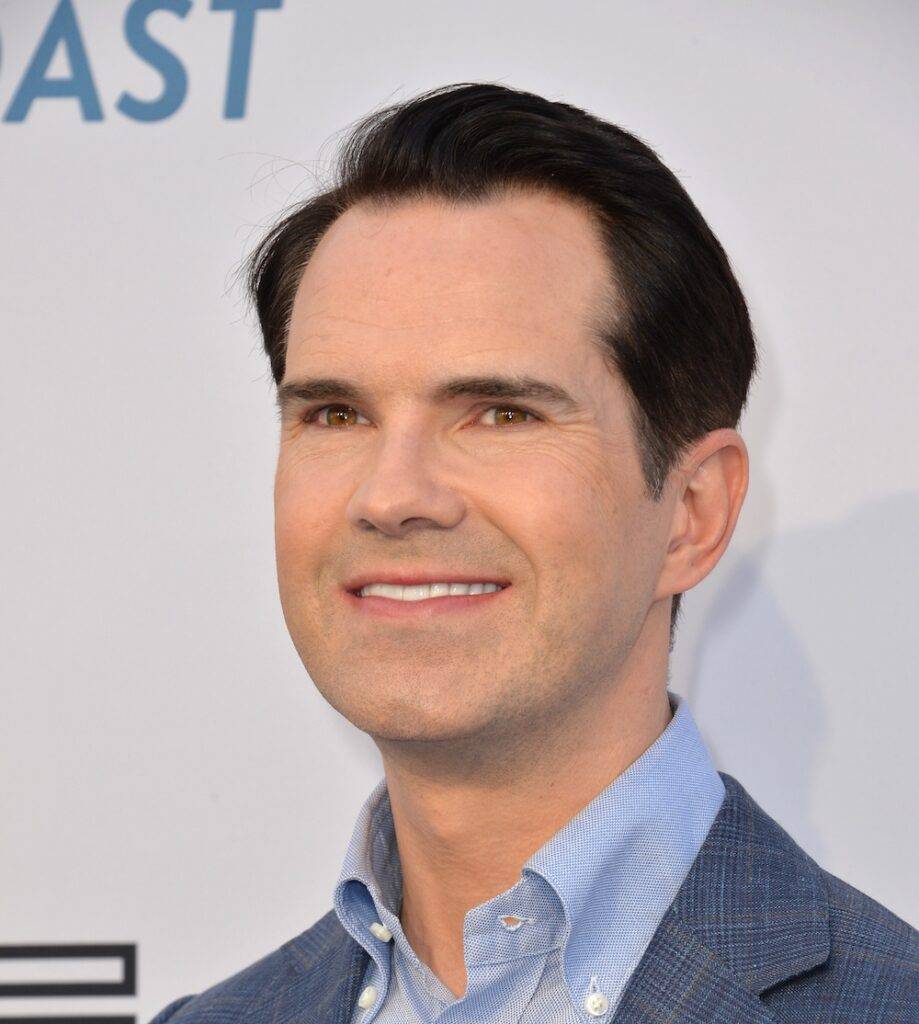 A middle-aged Caucasian man with signs of hair regrowth in his dark hair, smiling at a public event. He wears a light blue dress shirt under a textured blue blazer. The background includes a