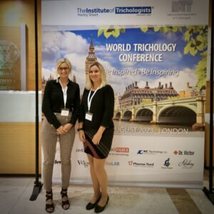 Two women in business attire standing in front of a conference banner that reads "world trichology conference" with images of London landmarks and hair replacement techniques in the background.