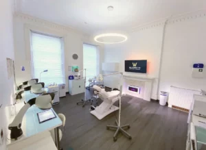 A modern dental clinic with two dental chairs, bright lighting, medical equipment, and a mounted TV displaying a clinic logo on a clean, white interior backdrop.