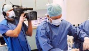 A videographer in protective gear films a surgeon performing a hair implant procedure in an operating room, capturing the medical process for educational or documentary purposes.