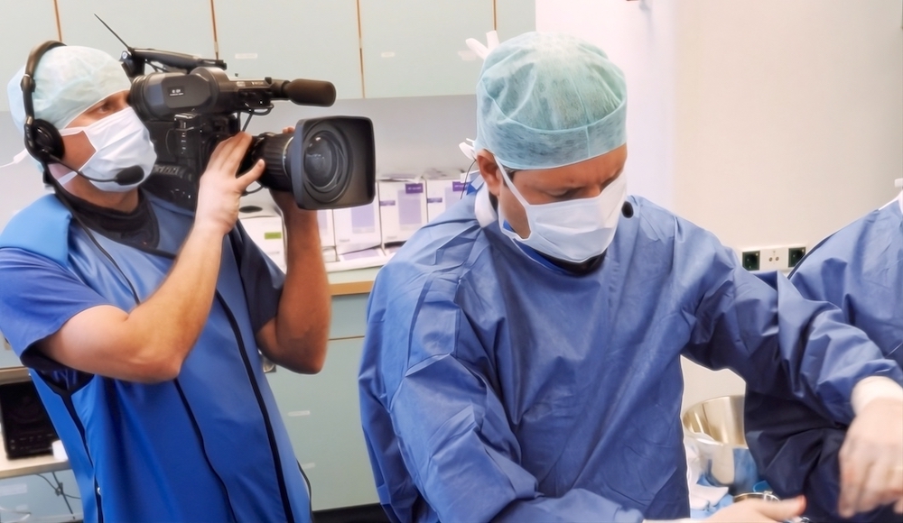 A videographer in protective gear films a surgeon performing a hair implant procedure in an operating room, capturing the medical process for educational or documentary purposes.