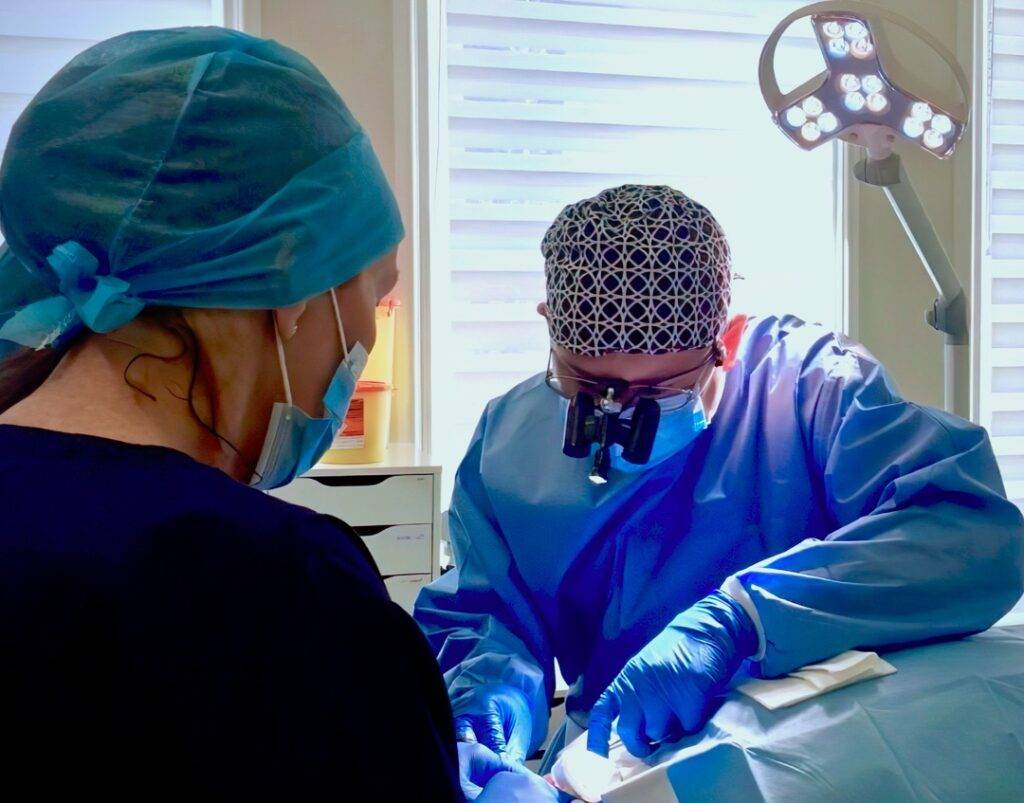 A surgeon in blue scrubs and a surgical cap uses a microscope and tools during a hair surgery procedure, with an assistant nearby under bright surgical lights.