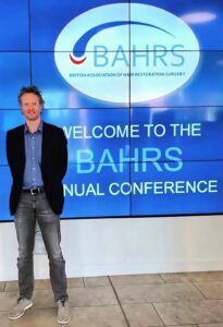 A man stands smiling in front of a sign reading "welcome to the BAHRs annual conference" at the British Association of Hair Restoration Surgery event focusing on hair implant.
