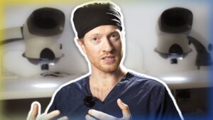 A man in blue medical scrubs and a black head cover speaks expressively about hair surgery, with surgical robotic arms blurred in the background.