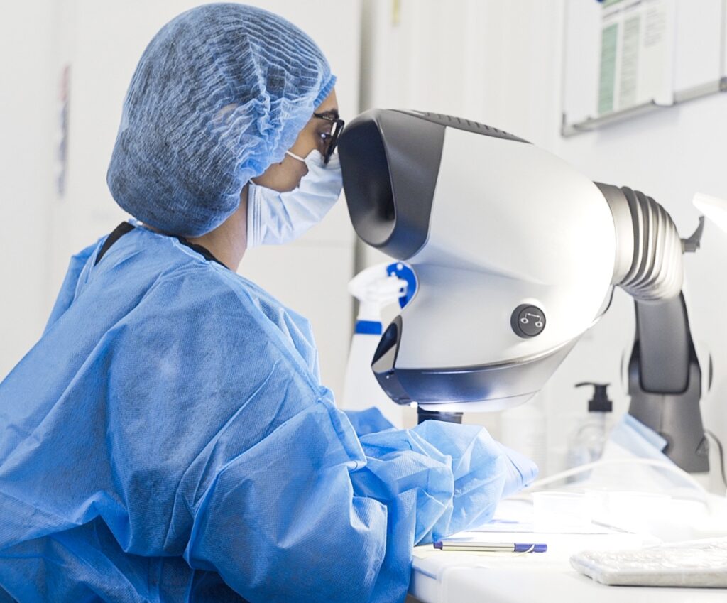 A female scientist wearing a surgical cap, mask, and blue scrubs closely examines a sample under a large, sophisticated microscope in a well-lit lab setting. This examination is fundamentally related to her research