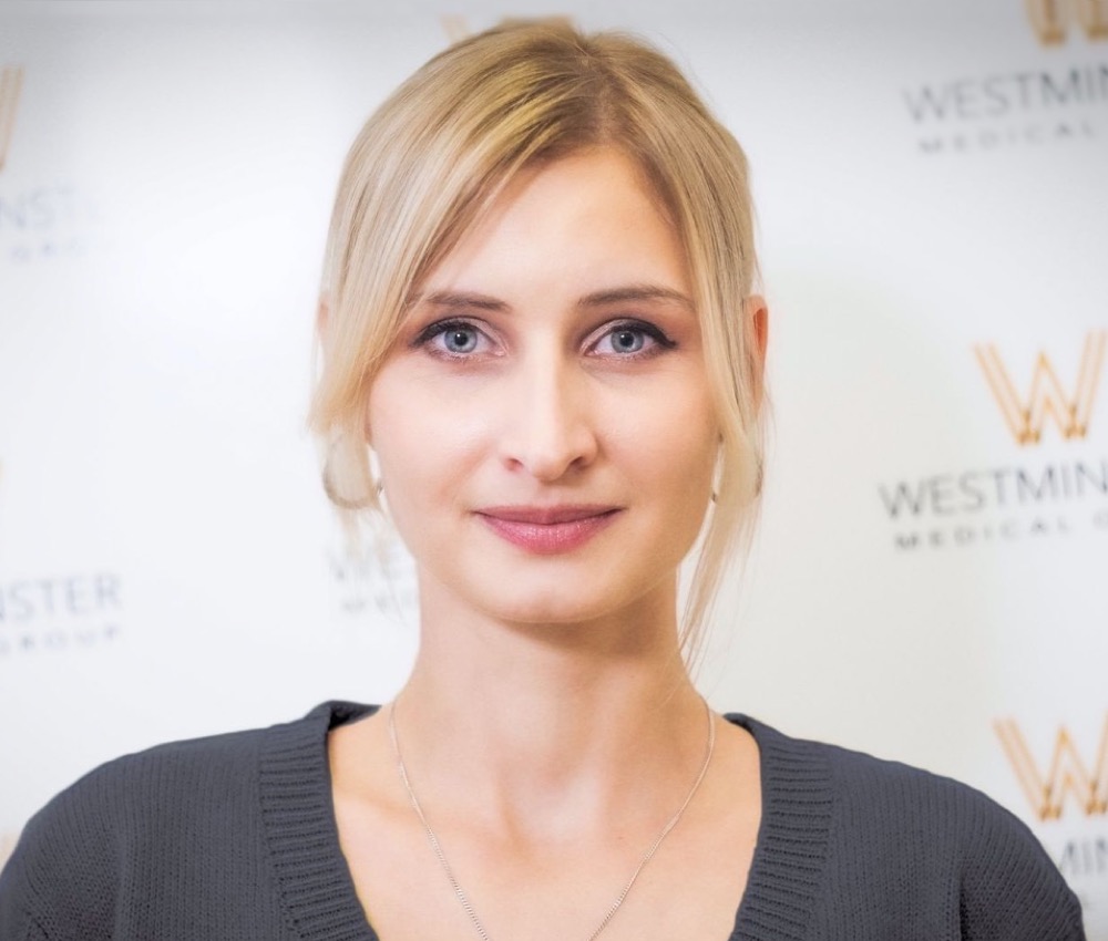 A woman with blonde hair and blue eyes smiling gently, wearing a dark sweater, standing in front of a backdrop with Westminster Medical Clinic logos specializing in female hair loss.