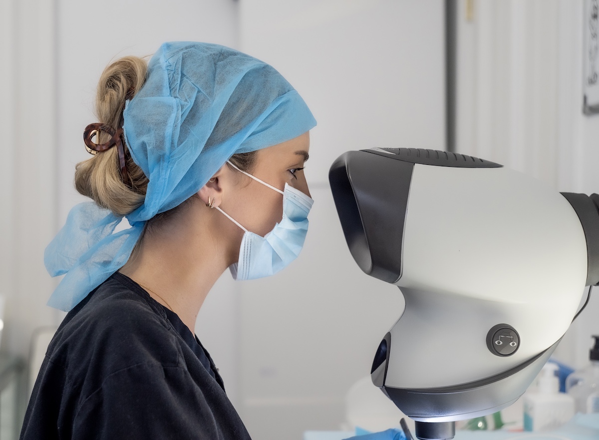 A female healthcare professional wearing a surgical cap and mask looks intently at a large, modern medical device for hair replacement in a clinical setting.