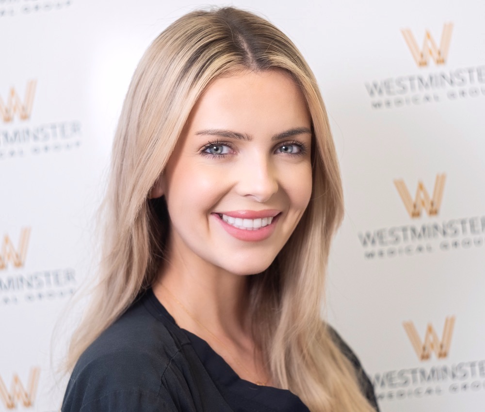 A smiling woman with long blonde hair poses for a portrait in front of a backdrop with the logo of Westminster Medical Group, specialists in hair replacement. She's wearing a black blouse.