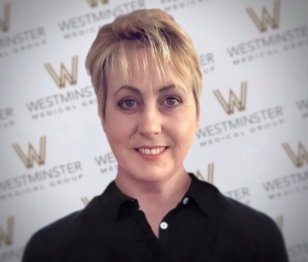 A portrait of a smiling woman with short blonde hair, wearing a black shirt, standing against a backdrop with the 'Westminster Medical Group' logo repeated, specializing in hair regrowth.