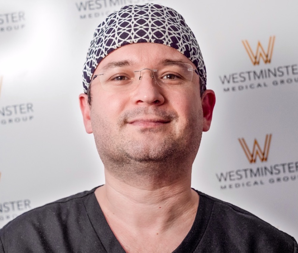 A smiling male healthcare professional wearing glasses and a patterned scrub cap stands in front of a backdrop with the Westminster Medical Group logo, specializing in male pattern baldness.