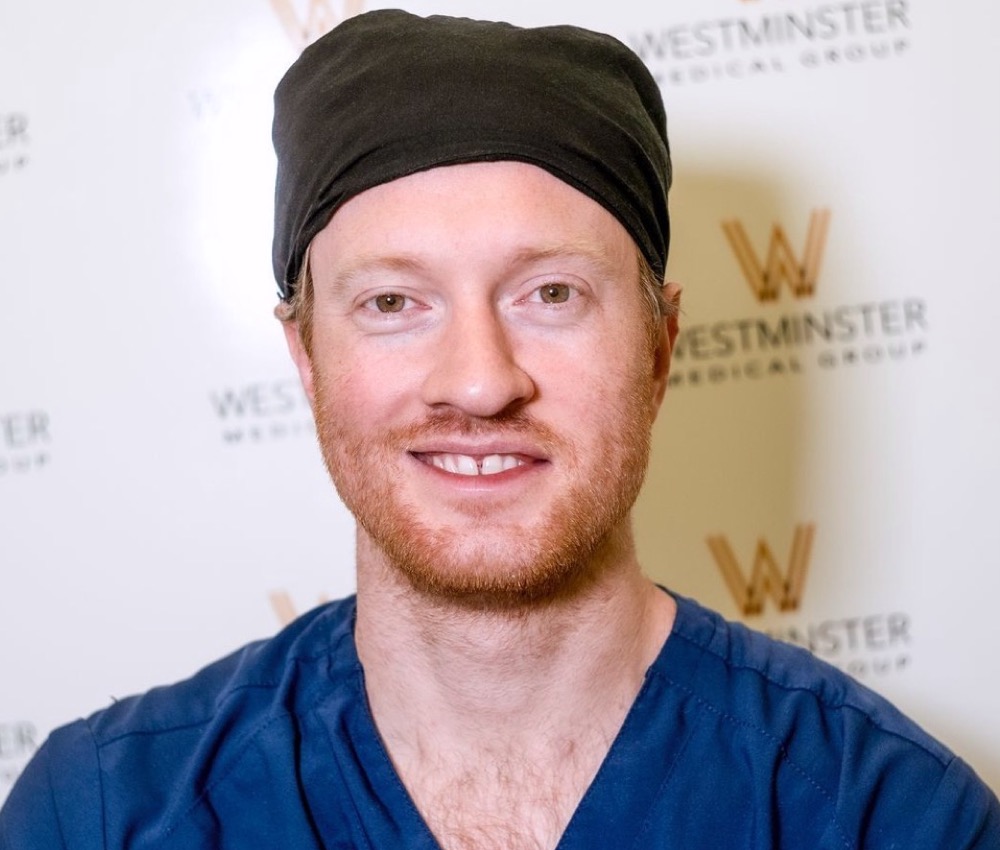 A smiling man wearing medical scrubs and a black head covering stands in front of a backdrop with the Westminster Medical Group logo, specializing in hair surgery.
