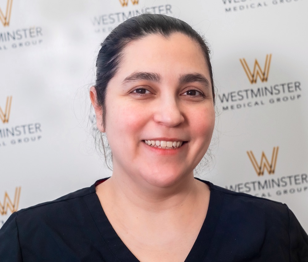 A woman with shoulder-length dark hair and a slight smile, wearing a dark medical scrub, stands in front of a backdrop with the Westminster Medical Group logo, specializing in female hair loss.
