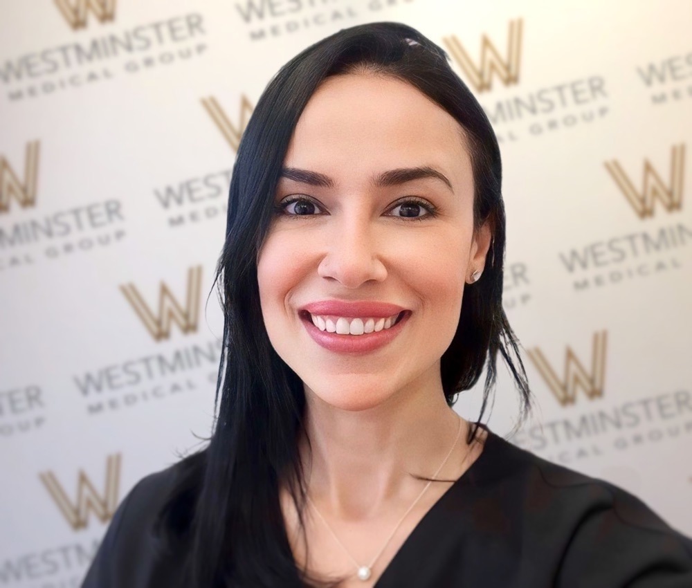A smiling woman with long dark hair, wearing a black top, stands in front of a backdrop with the "Westminster Medical Group" logo repeated, specializing in hair replacement.