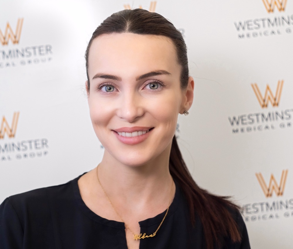 A professional portrait of a smiling woman with dark hair, wearing a black top, standing in front of a backdrop with the Westminster Medical Group logo, specializing in female hair loss.