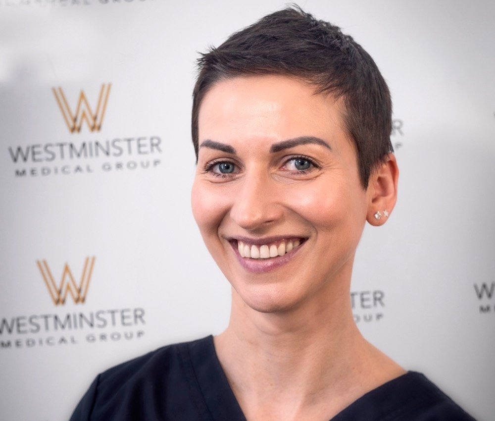A smiling woman with a pixie haircut, treated for female hair loss, wearing subtle makeup and small earrings, standing in front of a backdrop featuring the Westminster Medical Group logo.