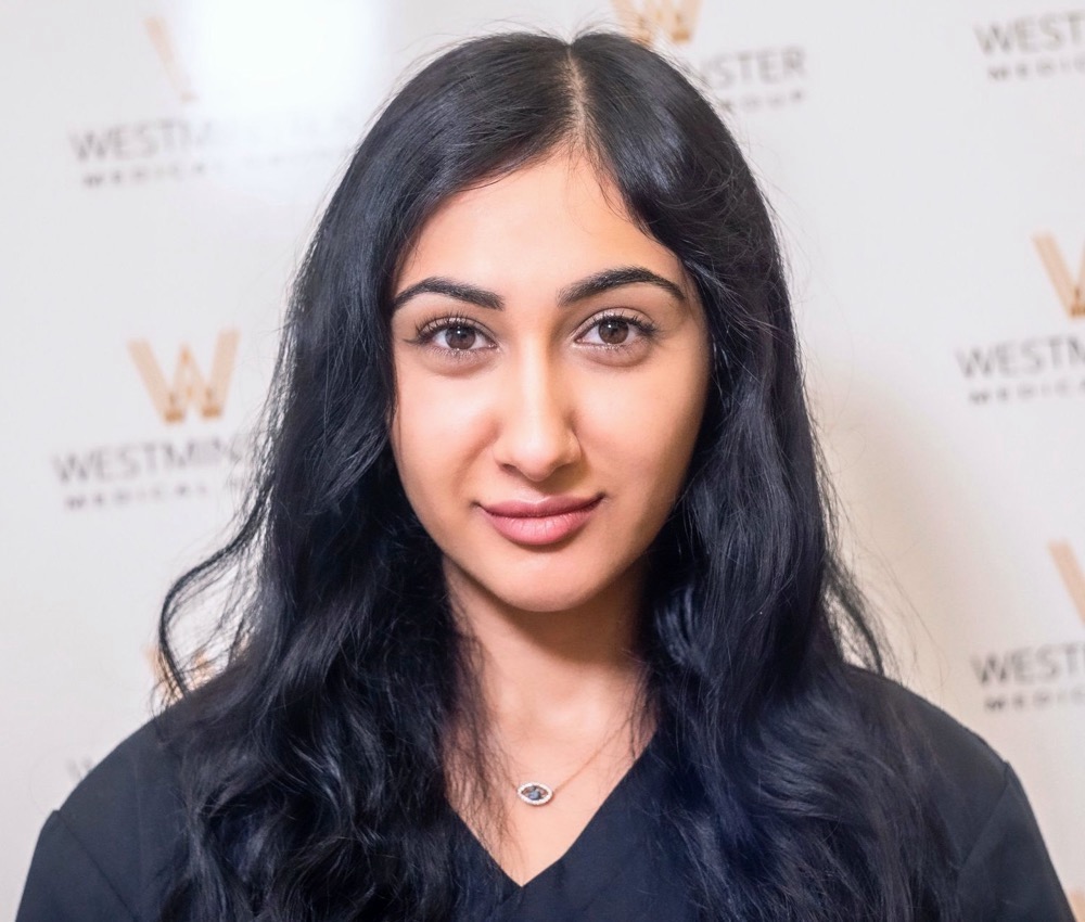 A young woman with signs of hair regrowth in her long black hair and dark eyes, smiling gently at the camera, with a background featuring logos on a beige wall.