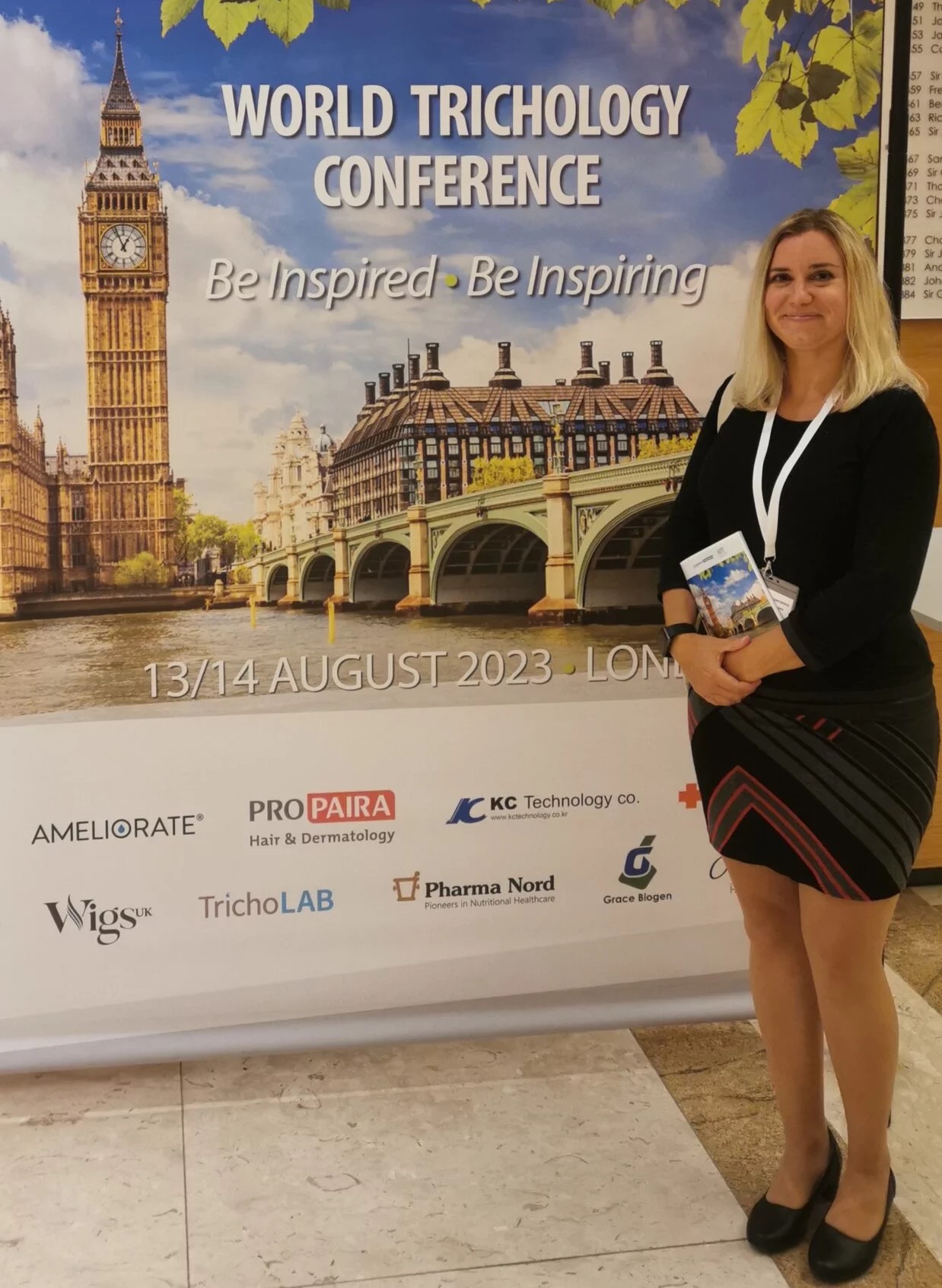 A woman stands smiling in front of a conference banner reading "World Trichology Conference" with an image of London landmarks. She wears a black outfit and holds a pamphlet on hair replacement.