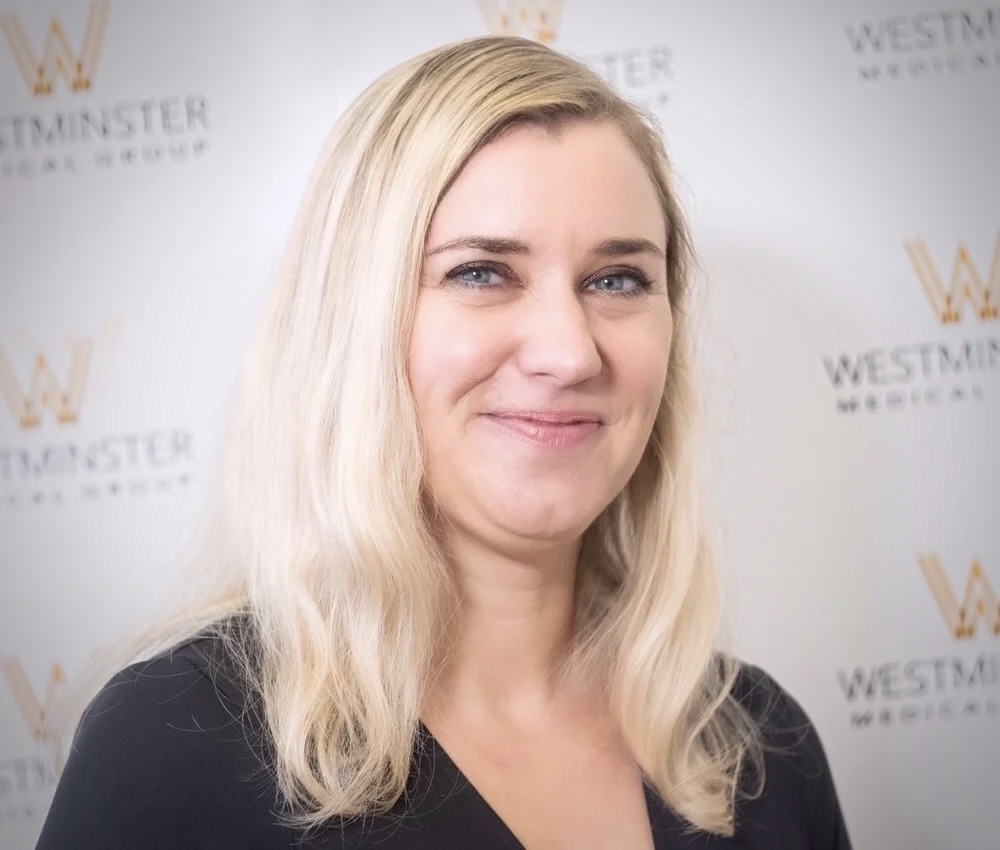 A smiling woman with blonde hair standing in front of a backdrop with the "Westminster Medical Group" logo repeated across it, specializes in treatments for female hair loss.