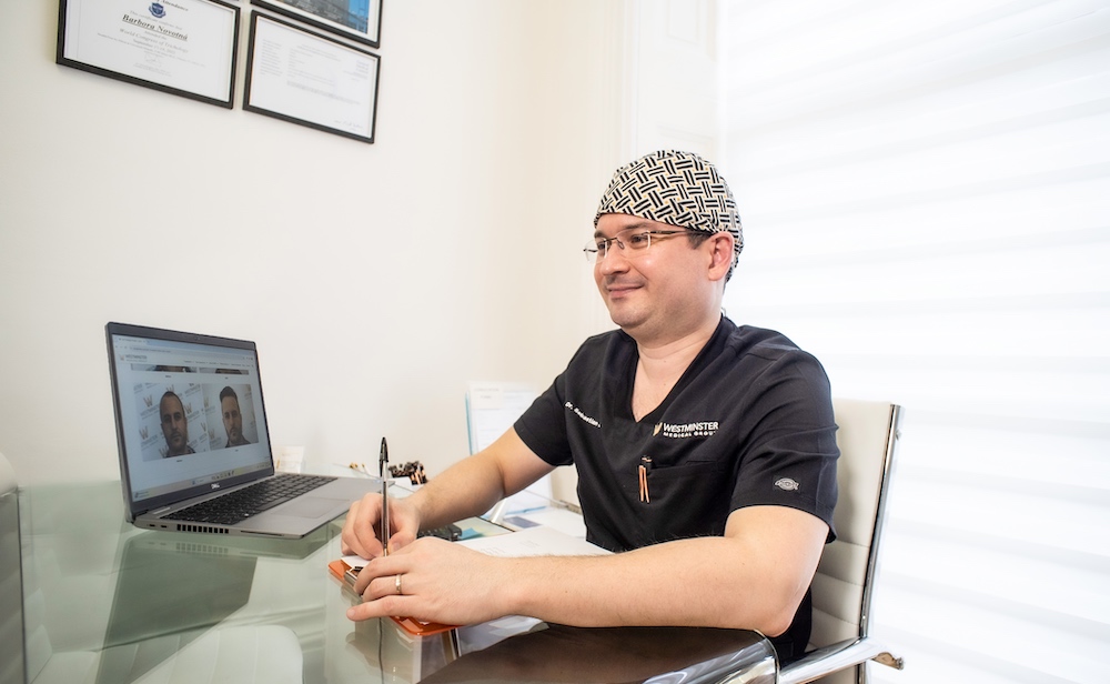 A male doctor in scrubs and a patterned surgical cap sits at a desk, smiling while looking at a laptop displaying images related to male pattern baldness, in a bright office with diplomas on