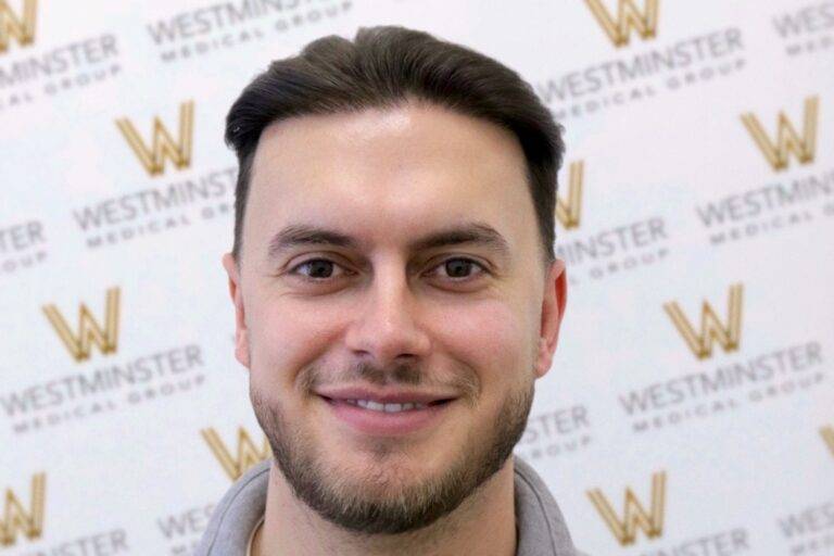 A smiling man with short dark hair in front of a backdrop featuring the Westminster Medical Group logo, specializing in hair surgery. He appears friendly and professional.