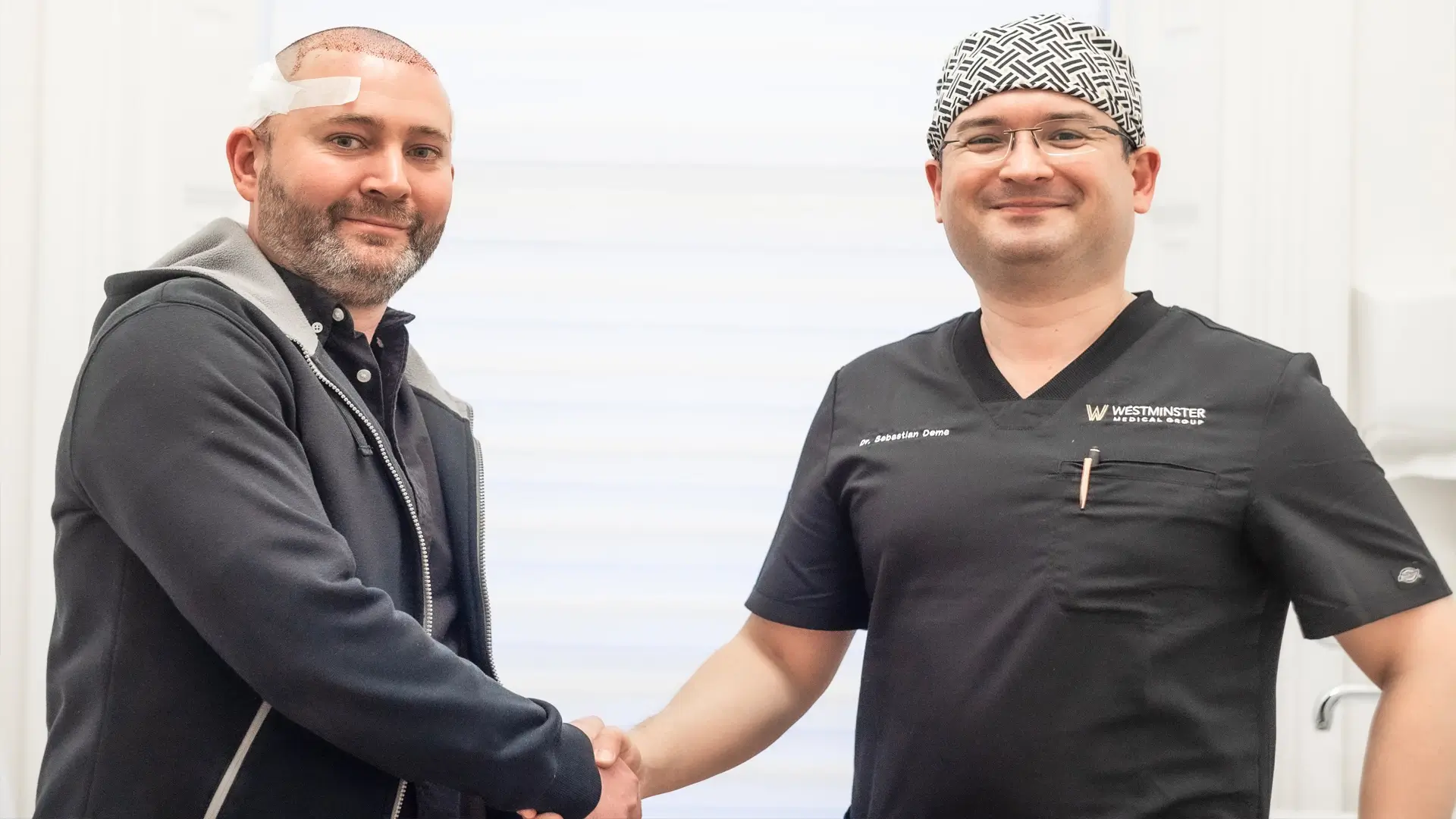 Two men shaking hands in a clinical setting; one in casual attire with a bandage on his forehead indicative of recent hair transplant services, and the other in medical scrubs and a patterned scrub cap