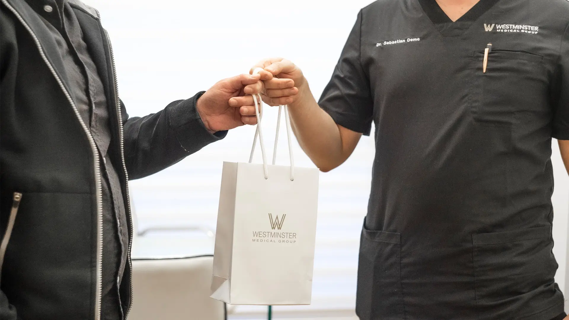 A person in a medical uniform hands a branded Westminster Medical Group bag, specializing in hair replacement, to another person, whose arm is visible. The setting appears professional, possibly within a clinic.